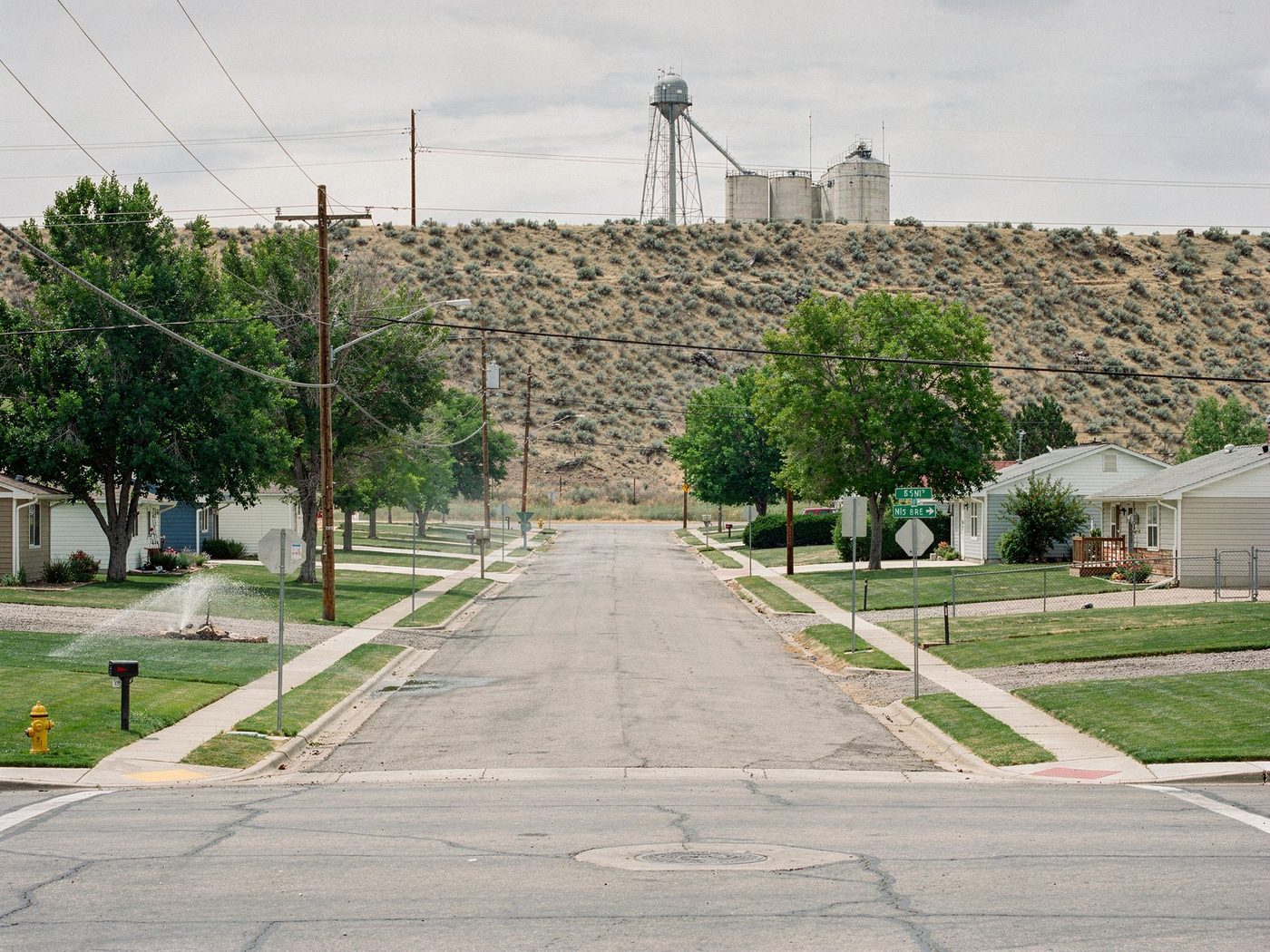 A residential street in an irrigation-project farm town at midsummer: watered lawns and shade trees, a sprinkler running, the town’s water tower and grain silos on the dry sagebrush bench above.