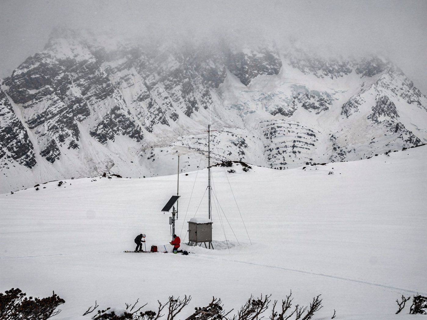 Two snow surveyors at a high telemetry station in falling snow, one probing the pack on skis, one kneeling at the instrument shelter with the record, storm light on the peaks behind.