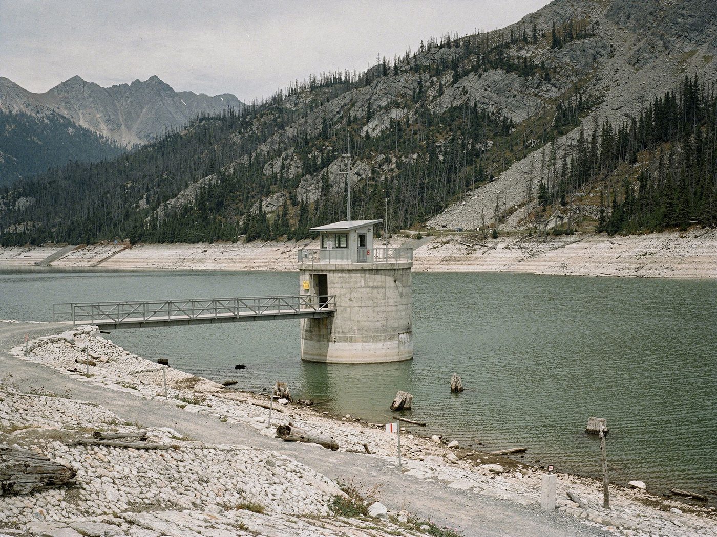 A reservoir outlet tower and its steel catwalk at autumn drawdown, the shoreline ringed by old water lines, drowned stumps standing clear of the pool, storm-killed timber on the slopes above.
