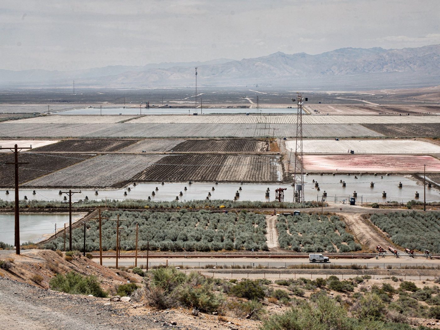 A vast dry alkali lakebed divided into an engineered grid of dust-control cells, some flooded, some dry, one stained pink, with power poles and a utility truck on the shore road.