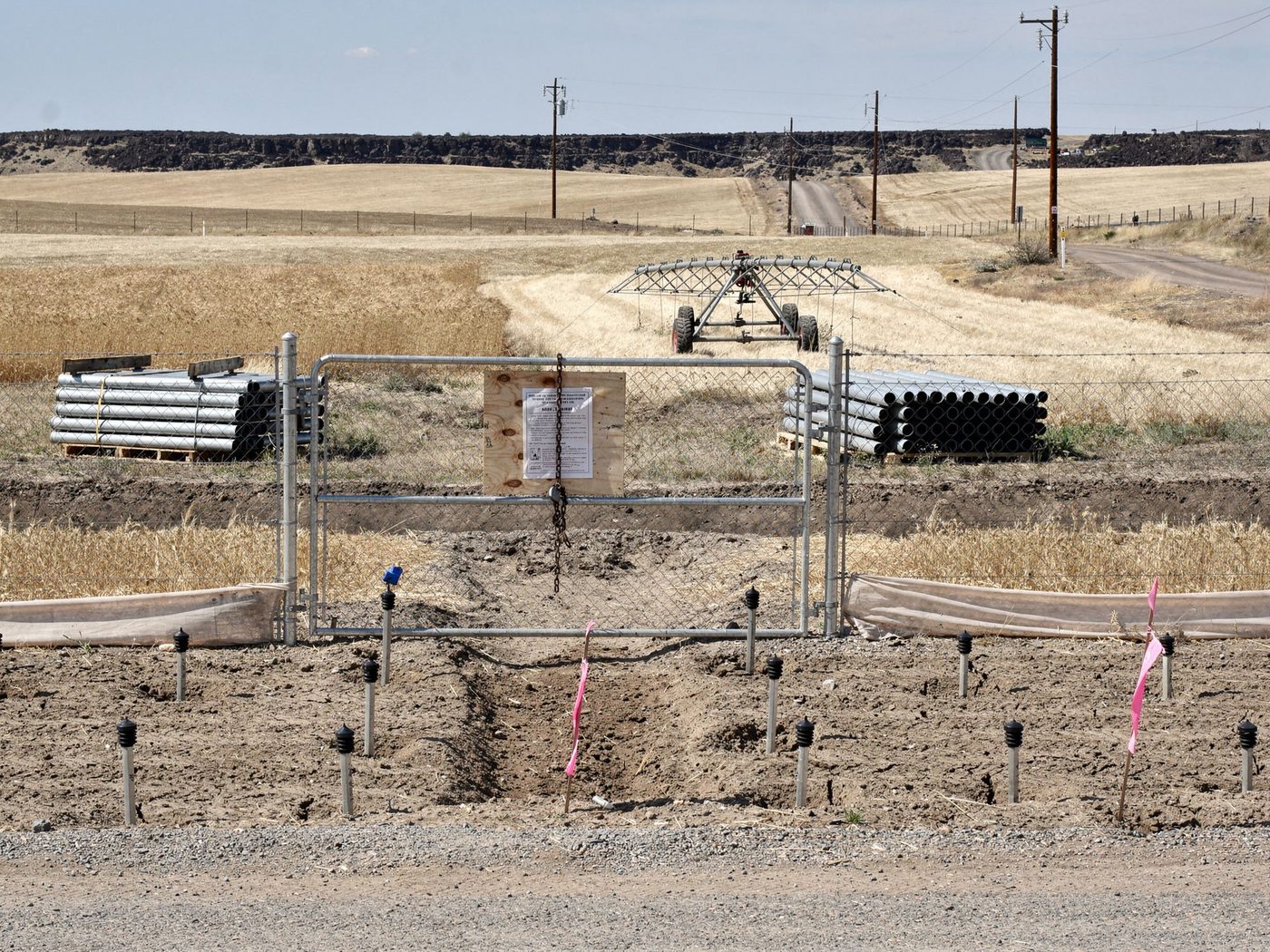 A chained gate with a posted public notice at a graded project site: survey stakes capped and flagged, silt fence run out, irrigation pipe stacked on pallets, and an idle pivot section parked in ripe wheat.