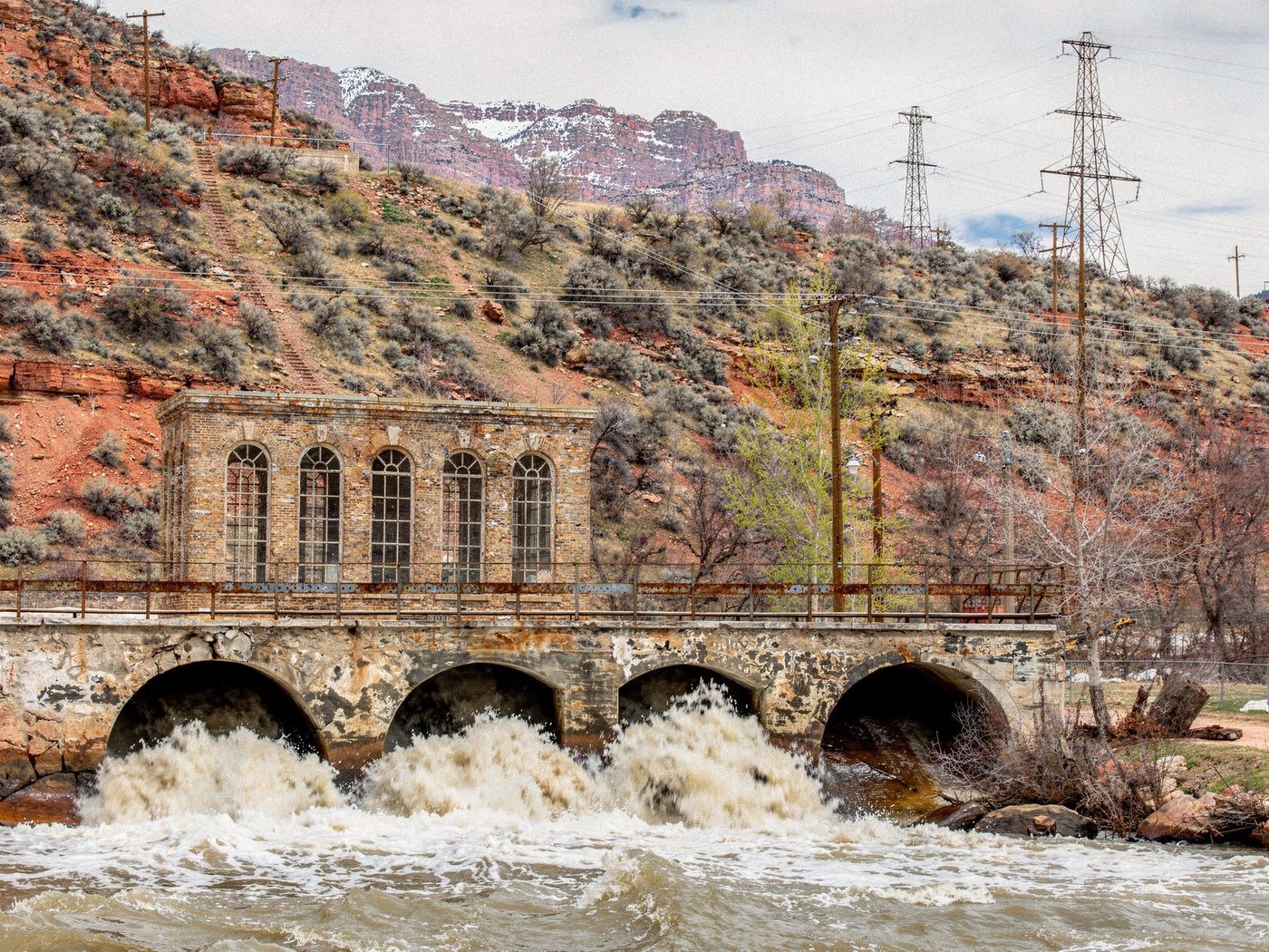 A century-old brick gatehouse above a four-arched river diversion at spring flood, muddy snowmelt driving through the arches, red-rock canyon walls and transmission towers behind.