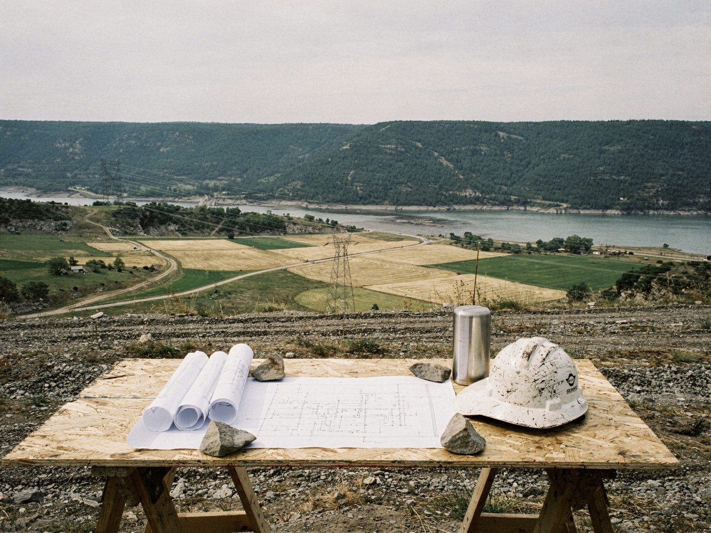 A plans table at the rim of a graded bench — rolled drawings and an open sheet weighted with stones on plywood over sawhorses, a worn hard hat and steel flask beside them — above a valley of cut and green fields, a transmission line crossing toward a reservoir under timbered slopes.