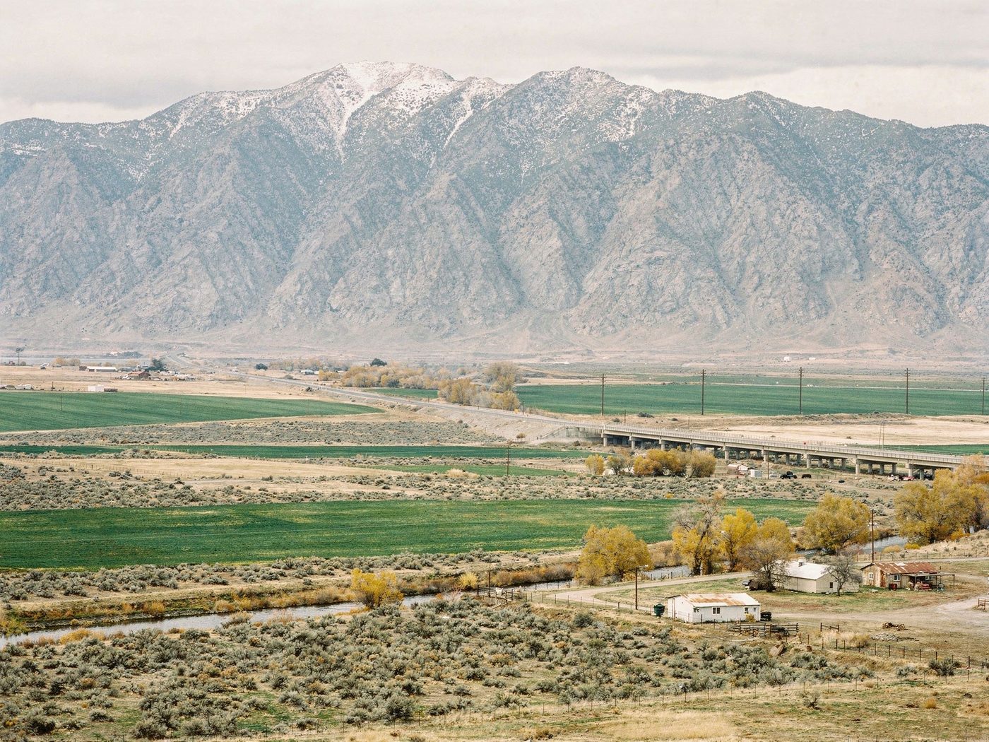 A working valley from above: a snow-dusted mountain front, green irrigated fields set against sagebrush, a river winding past small ranches, a highway crossing the bottomland on a long viaduct.