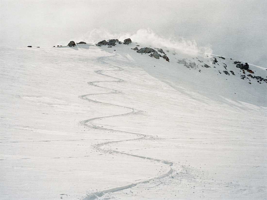 A single ski track descending an untracked snowfield below a wind-scoured ridge, spindrift blowing off the crest.
