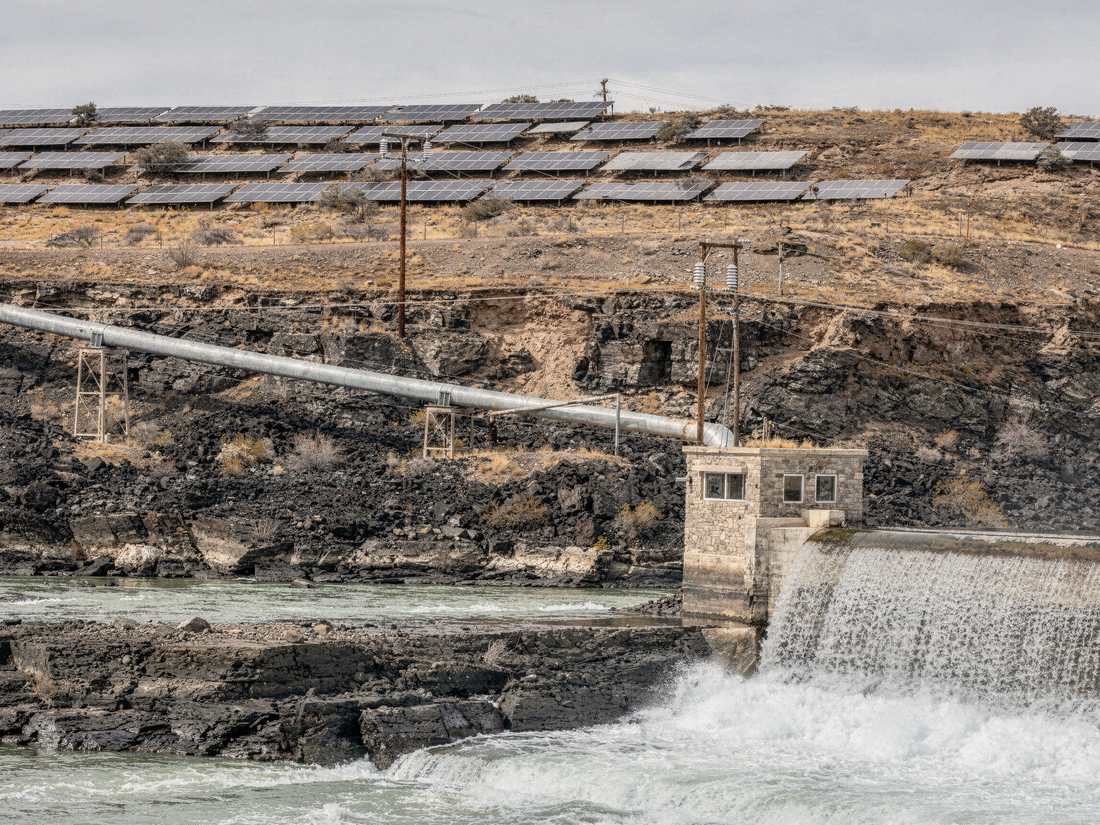 A diversion dam spilling into a basalt gorge, a penstock running out along the rim, and a solar array laid across the bench above it.