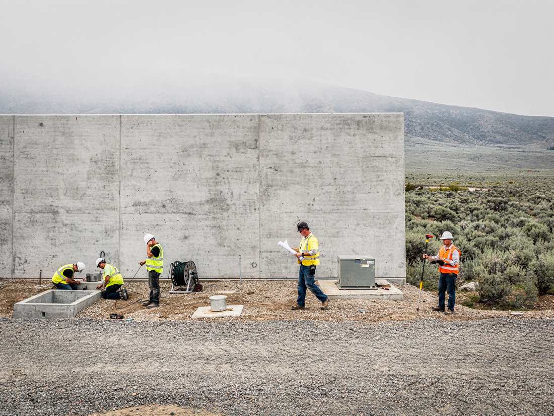 A crew working a row of ground fixtures along a blank tilt-up concrete wall where graded gravel meets unbroken sagebrush, hills fading in haze behind.