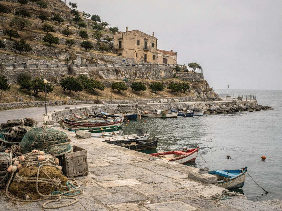 A working harbour on a terraced coast: open boats made fast along a stone quay, nets and floats stacked on the flagstones, lemon groves stepping up the slope behind.