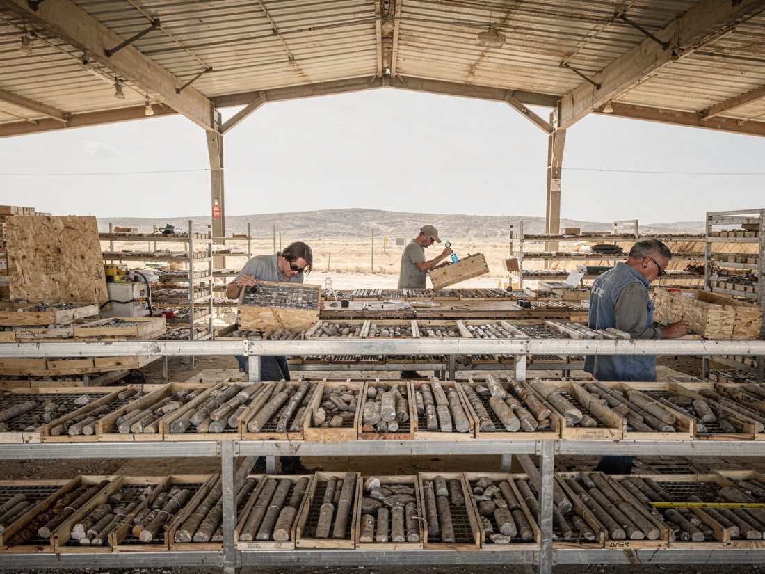 Three geologists logging drill core at long racks under an open shed roof, desert bench and haze through the far opening.