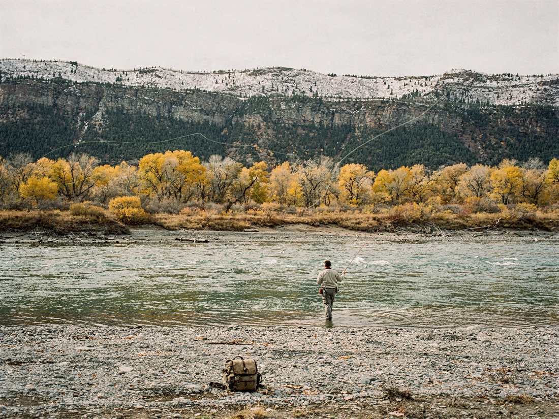 A single figure fishing a wide river, a pack set on the gravel bar in the foreground, cottonwoods in autumn and snow-dusted rimrock behind.